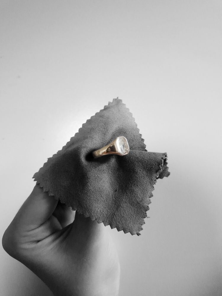 Black and white photo of a hand holding a signet ring with a polishing cloth. The black and white filter covers everything apart from the ring, which is gold and highly polished. 