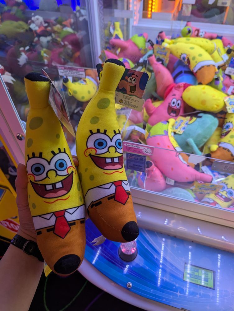 holding two banana-shaped SpongeBob plushies while standing in front of a crane machine filled with various plushies of SpongeBob characters