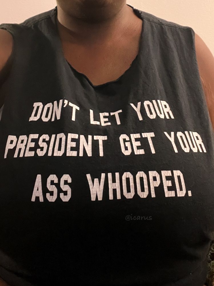 Image of me, wearing a black t shirt that I cut the sleeves and neck off of. It has white text that reads “Don’t let your president get your ass whooped”