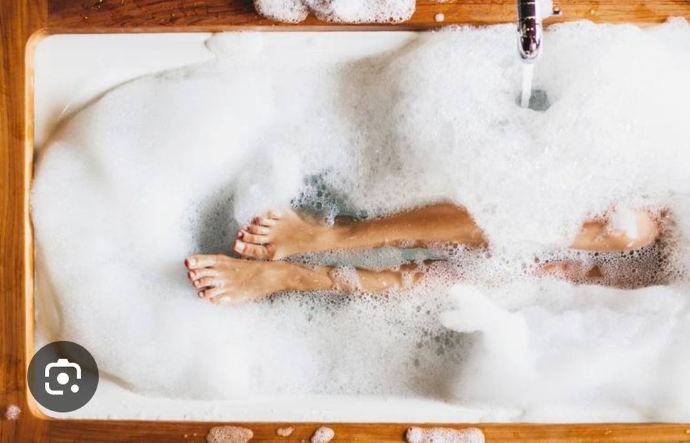 Badewanne gefüllt mit Wasser und Schsum von oben zu sehen. Beine und Füße sind zu sehen.