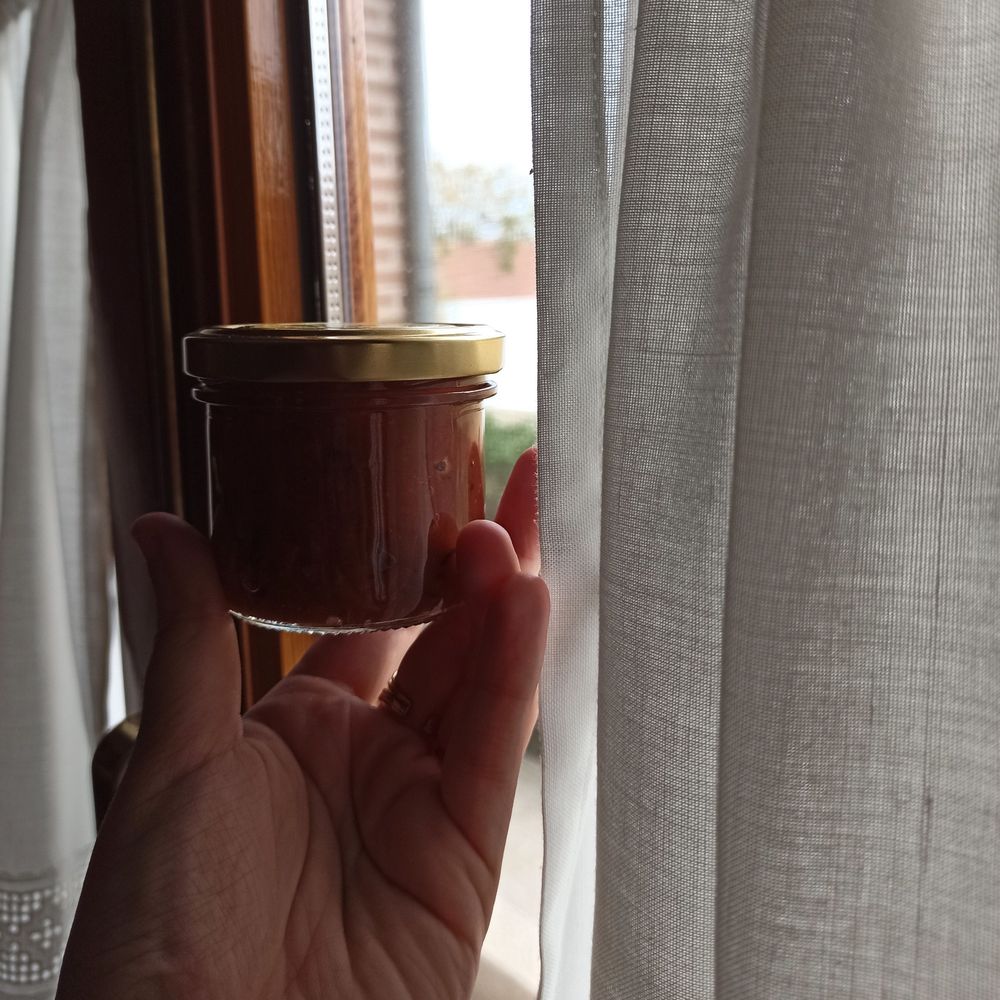 Bote pequeño sujetado con la mano al contraluz en la ventana. Las cortinas son blancas, la ventana de madera, el bote contiene chutney de color caramelo y la tapa es dorada.