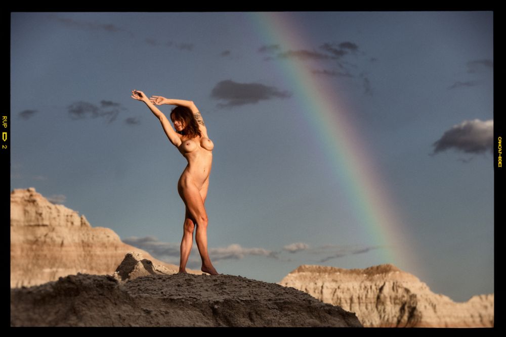 A nude woman stands on a rocky outcrop in the foreground. Badlands buttes rise behind her. The grey-blue sky is punctuated by a few clouds and a rainbow. 