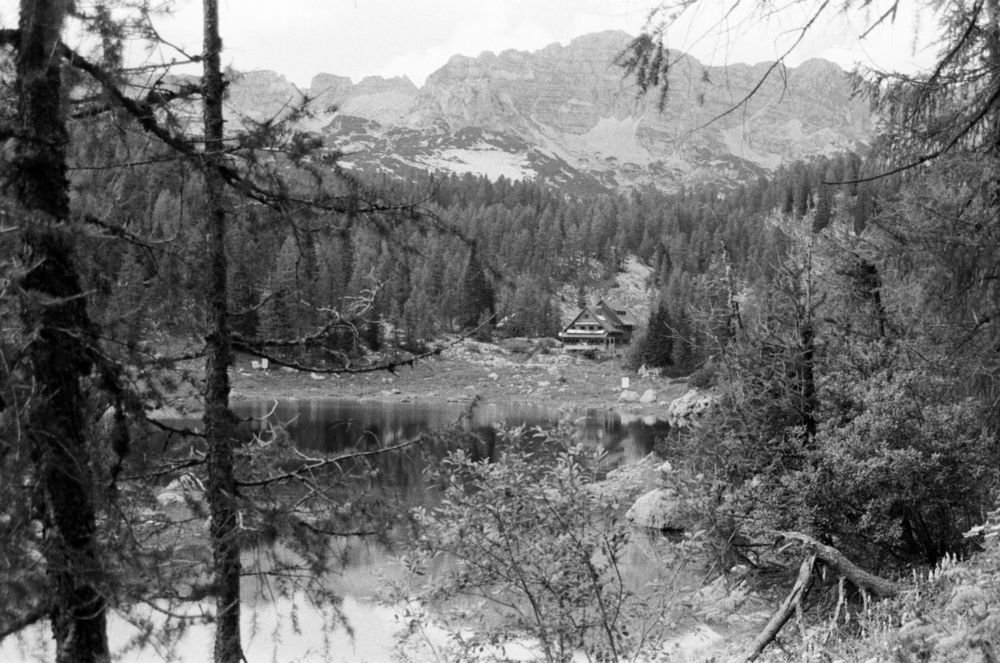 A black and white photograph of an Alpine hut behind a lake. There is a forest behind the hut, and mountains in the background, as well as some trees in the foreground.