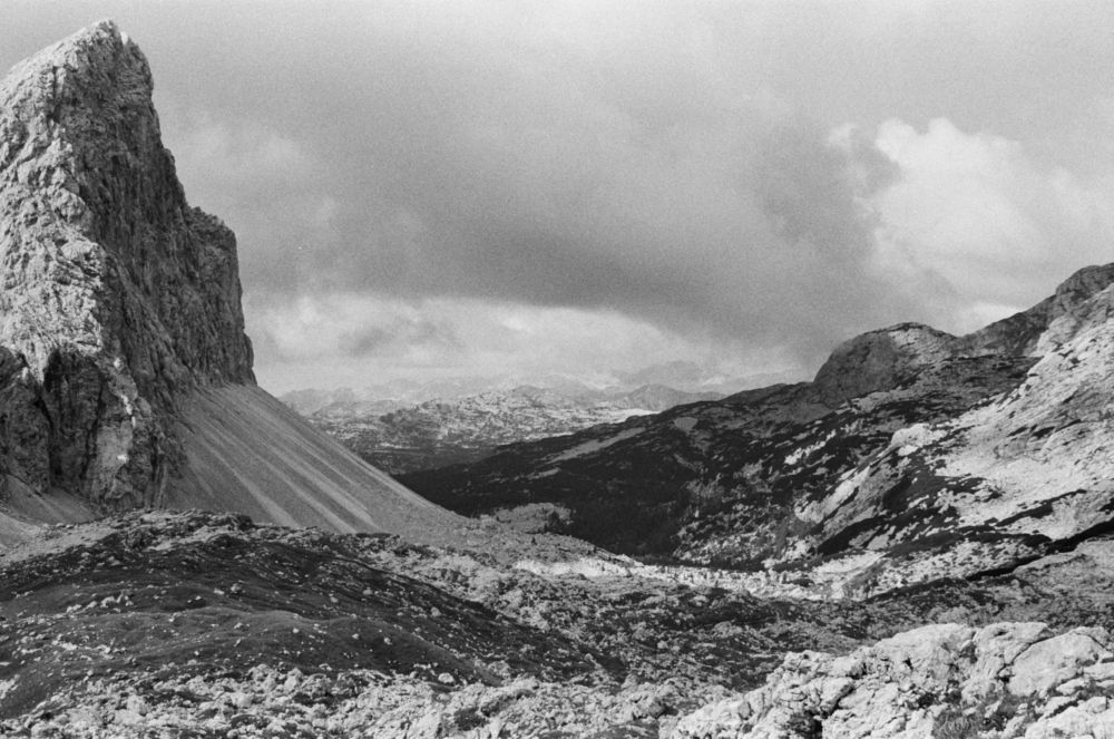 A black and white photograph looking down a valley in a mountainous area. There is a peak in the left of the image, with a scree slope below it, and a gentler slope on the opposite side of the valley. A forest is visible in the distance further down the valley. Other mountains can be seen in the far background.
