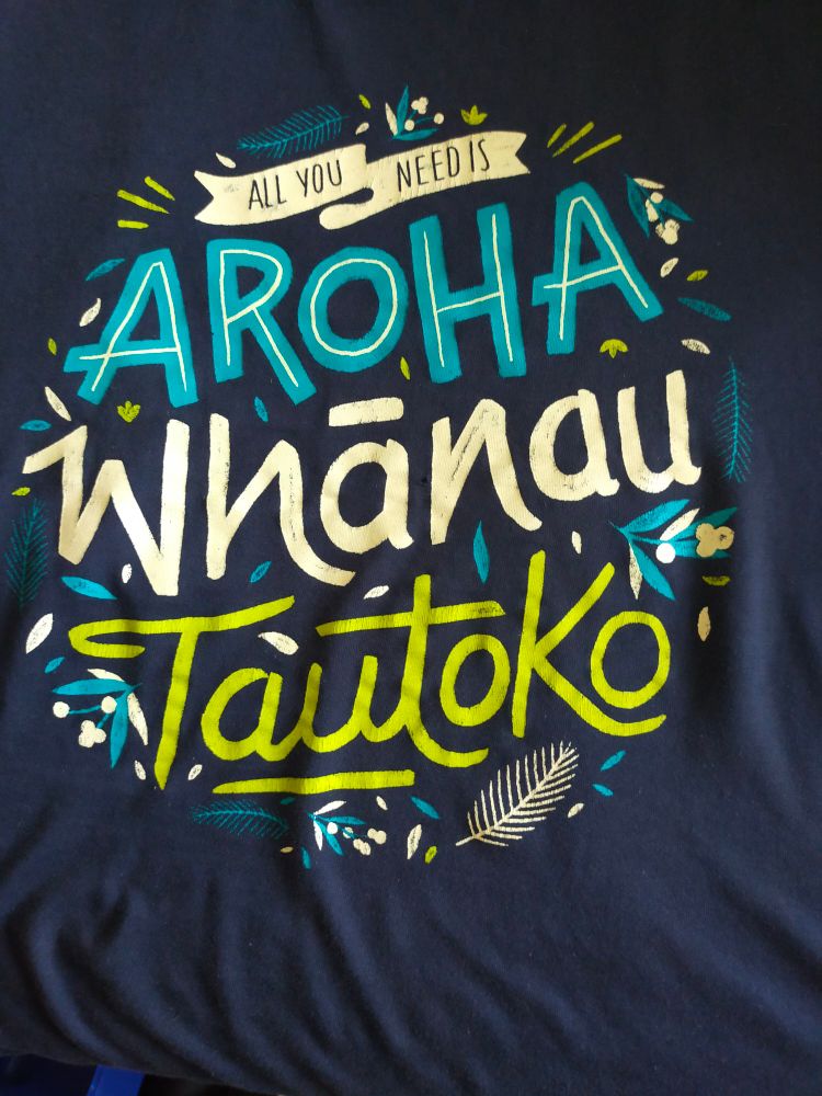 Blue t-shirt with lighter block lettering that reads "all you need is aroha, whānau, tautoko!"