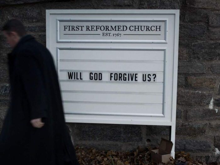 A priest walks past a church sign that reads WILL GOD FORGIVE US? From First Reformed (2017).
