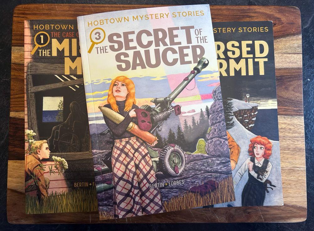 Three books are stacked on a wooden cutting board. Obscured on the bottom row are Hobtown Mystery Stories Volumes 1 and 2, and on top in clear view is Hobtown Mystery Stories Volume 3: The Secret of the Saucer. On the cover a young woman with long blonde hair wearing a long tartan skirt looks up at the sky while holding an artillery shell and balaclava. A missile shooter is behind her, and peeking out by some rocks in the background is a green bigfoot-looking creature.