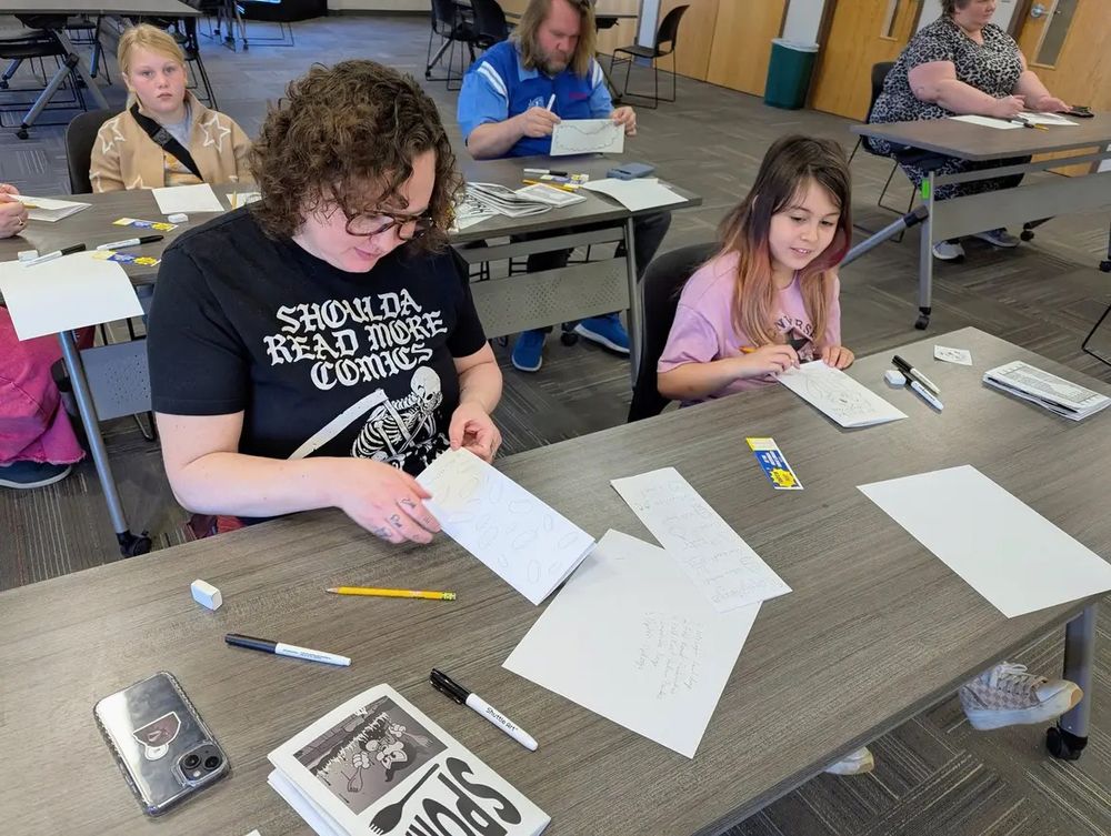 A woman and young girl sit at a table holding small hand-made comics they’ve just finished drawing.