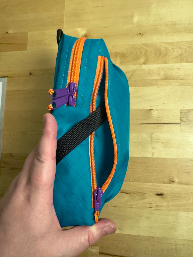 The side of a small bag showing the opening of an outer pocket. There is webbing coming out that leads to a key clip (not visible). The bag is teal with bright orange zippers and royal purple zipper sliders. 