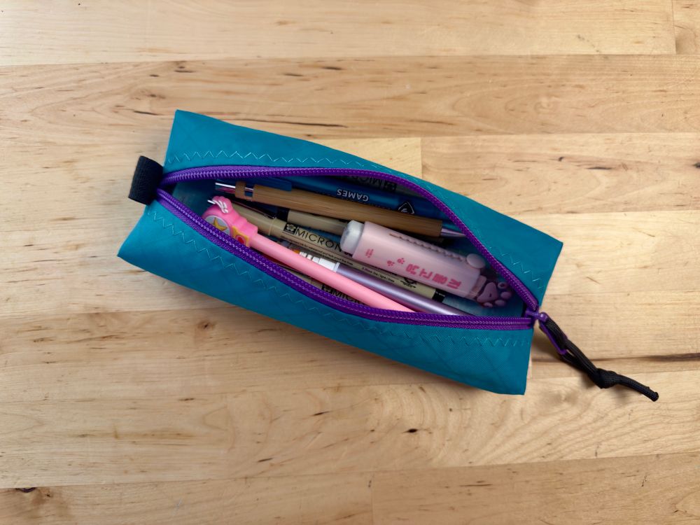 A teal pencil case with purple zipper, zig-zag top stitch and black paracord sipper pull. Opened showing several pens pencils and an eraser.  It is laying on a wooden surface. 