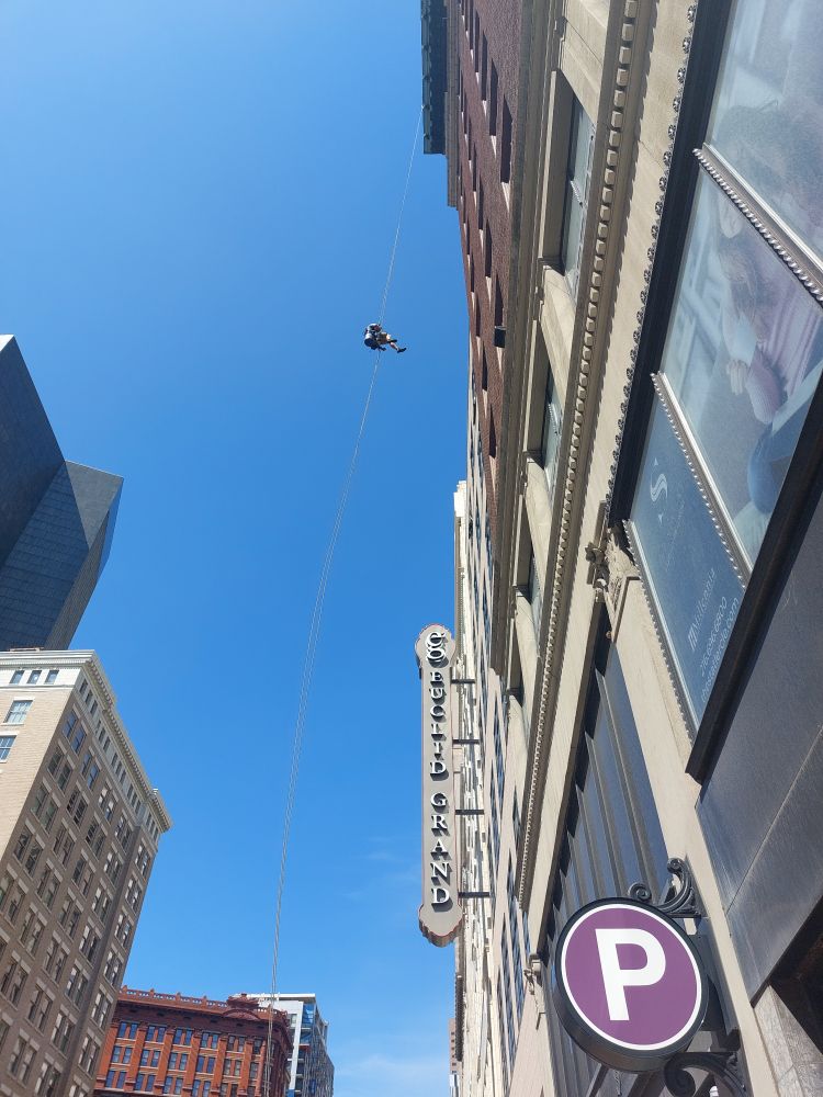 Somebody rappelling down the side of a downtown building, as seen by me from below. Nearly clear blue sky above