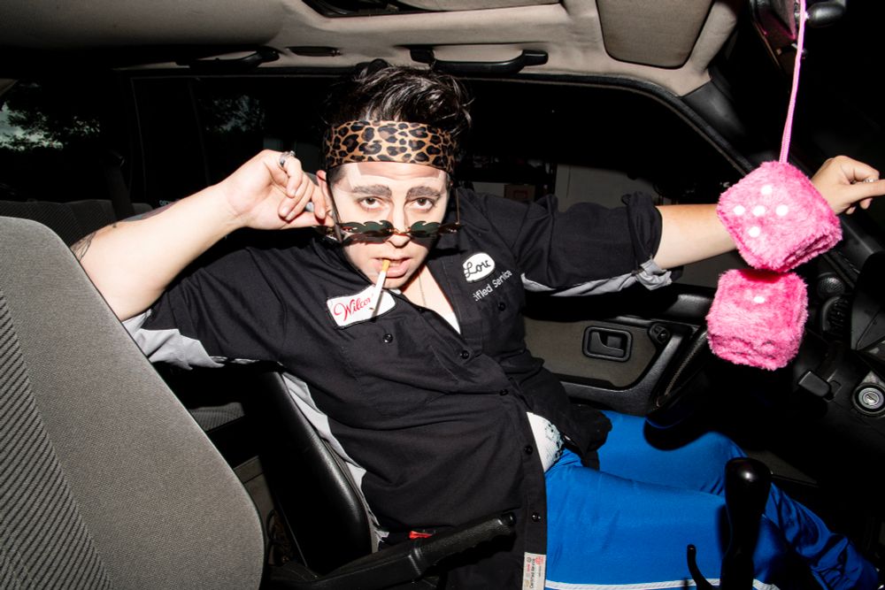 Portrait of a drag king wearing a mechanic’s shirt and leopard print head band sitting in the drivers seat of a vehicle, pulling down their glasses and smoking a cigarette. Pink fuzzy dice hang from the rear view window. 