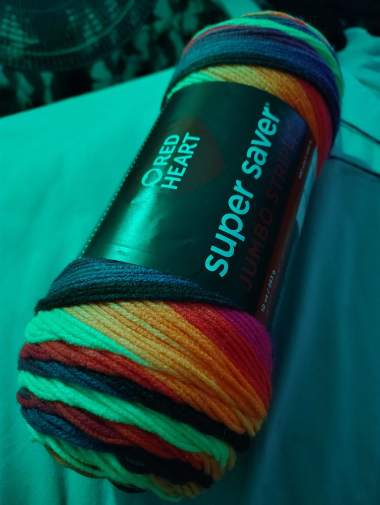 A skein of yarn - the yarn is neon rainbow and black, all fading into each other. It sits on the corner of the bed in blue light.