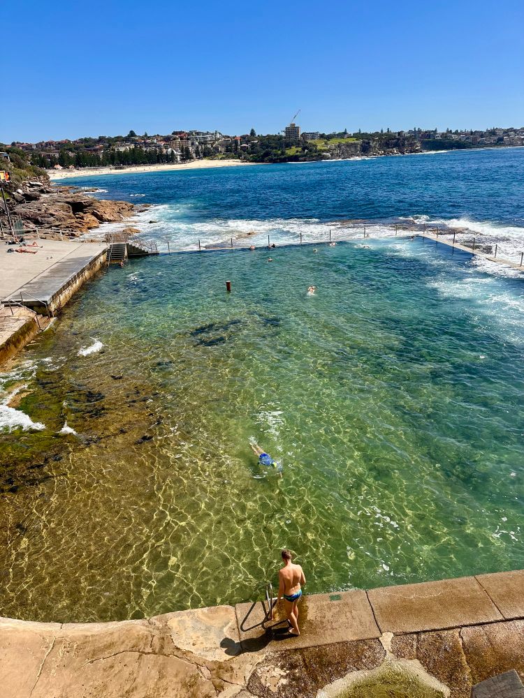 An ocean rock pool sparkling in the sunshine as several swimmers frolic in the crystal clear waters. In the background is a long beach & some houses
