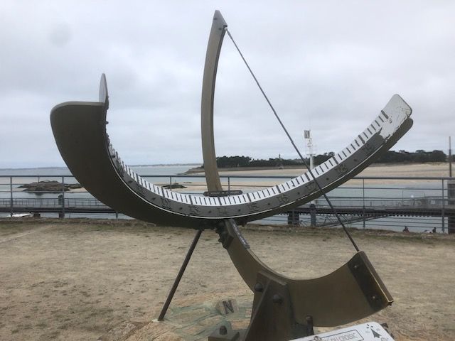 A metallic equatorial sundial, made of a concave band carrying hour lines, supported by a perpendicular meridian, and a thin rod casting its shadow. Photo taken on a cloudy day. Sea in the background.