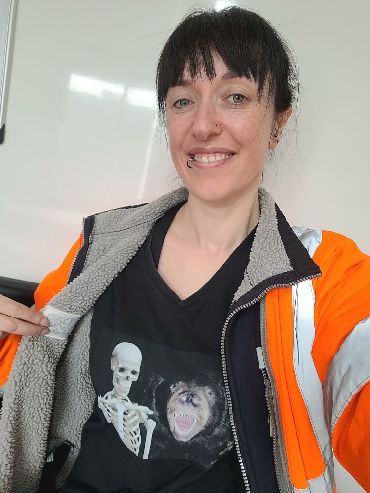 A selfie of a woman with dark hair wearing a orange high visibility best and dark blue fleece. She holds open the zip to reveal a black tshirt which has an image of a human skeleton and a bear with its mouth open