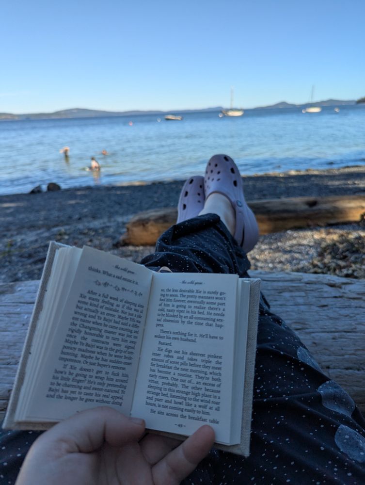 My hand holding a tiny book open. My legs are stretched out and you can see my lavender purple crocs and the ocean. 