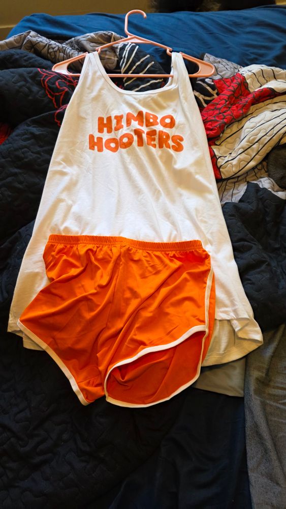 A white tank that says "Himbo Hooters" in the Hooters font and orange boots shorts. 