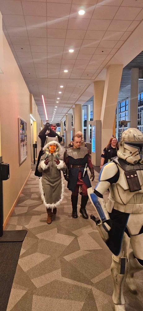 Captain Rex leads the way as Ahsoka and Anakin follow behind. Ahsoka holds up two peace signs with a smile while Baylan Skoll holds his lightsaber over Ahsoka’s head. A handler laughs off to the side. 
