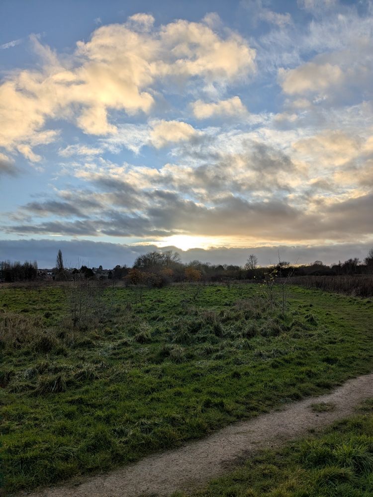 Photograph of grass and a path in the foreground with a mixture of clouds and blue sky, lit by a setting sun. Trees on the horizon.