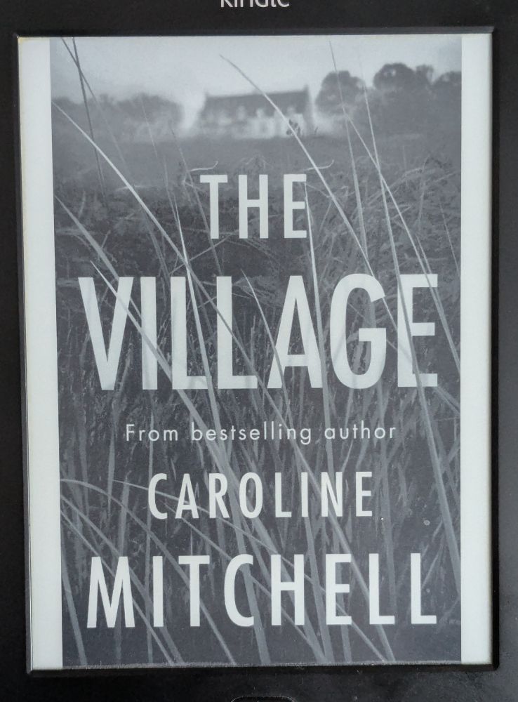 Kindle ebook cover of 'The Village' by Caroline Mitchell, featuring tall grass and houses and blurred trees in the distance.