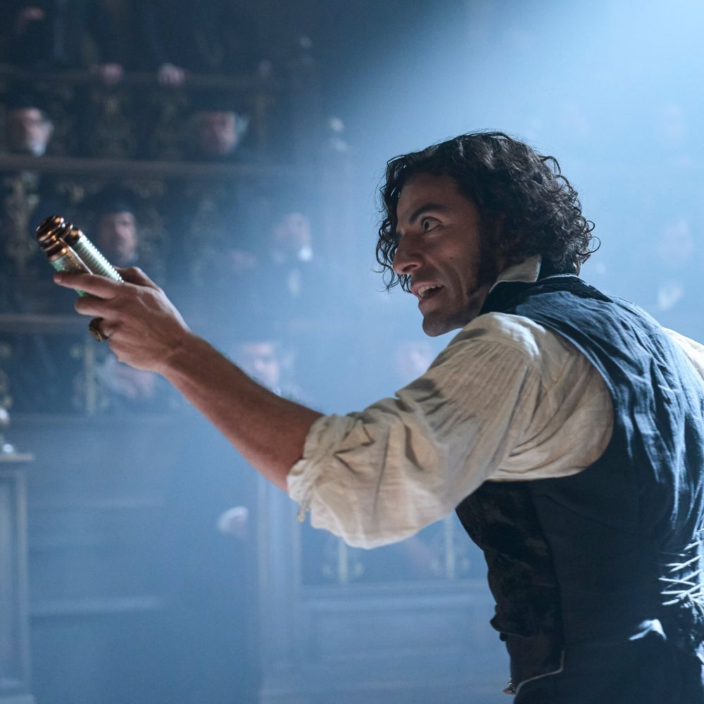 Oscar Isaac as Victor Frankenstein from the 2025 film, 'Frankenstein'. A man with dark curly hair in a white shirt and black waistcoat holds up a power cell to an auditorium with a shaft of light shining down on him as he speaks.