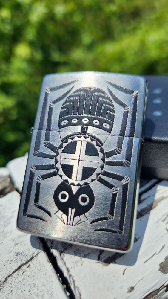 A photo of a brushed chrome zippo lighter with a printed black cherokee water spider design. The spider is facing downward with the fire, represented by a circle with a cross, on her back. 