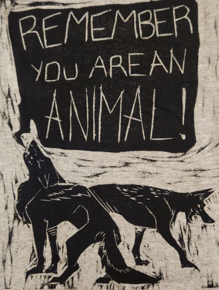 A woodcut print in black ink on grey fabric of a pair of cackling and howling coyotes. A big speech bubble above them reads ‘REMEMBER you are an ANIMAL!’  