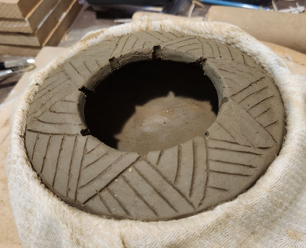 A clay cazuela bowl sitting inside a bowl mold lined with cloth. The rim is decorated with semicircular cutouts and the top has triangular patterns of parallel lines incised into it. The clay is still wet