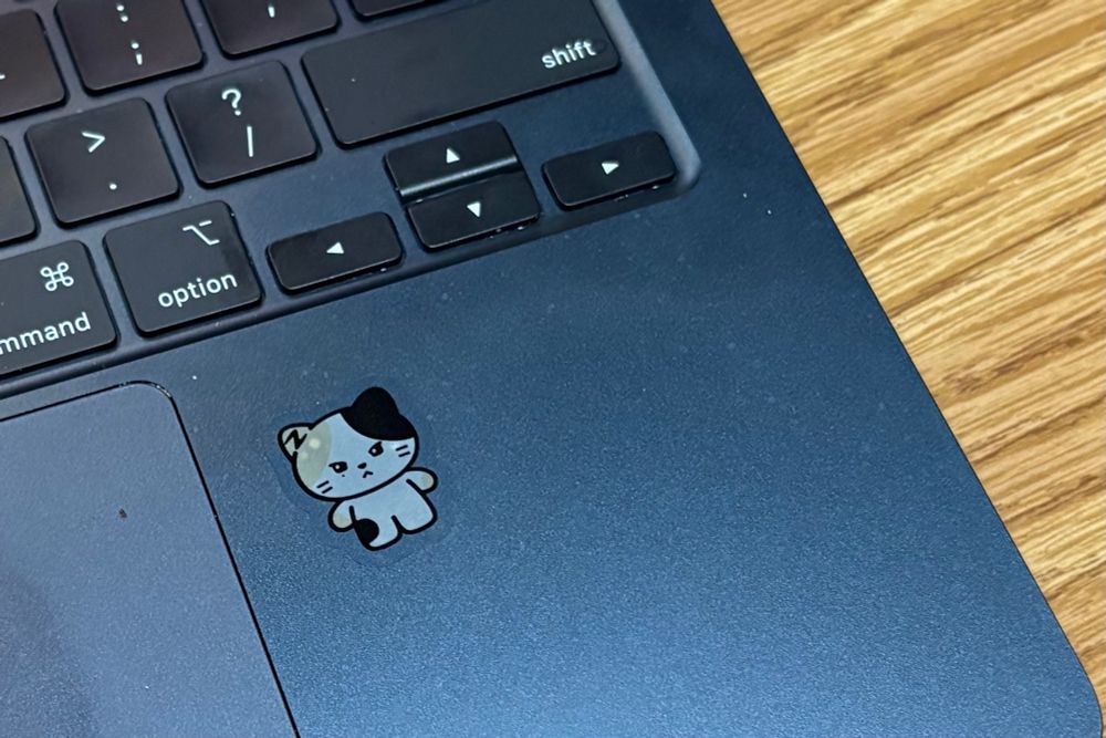 Photo of a sticker next to the track pad of a black laptop. The sticker is a drawing of Rinini, a cartoon calico cat character that represents Shen Ricky from the k-pop group Zerobaseone.
