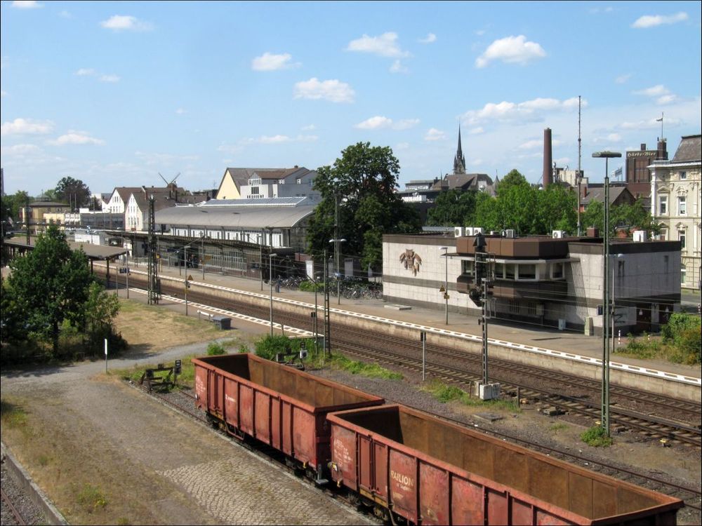 Blick aus erhöhter Position auf einen Bahnhof. Vorne zwei leere, rotbraune Güterwagen. Dahinter zwei Gleise mit Bahnsteigen. Hinter einem Signal das Stellwerk, links davon das Empfangsgebäude. Hinten u.a. Kirchturm und zwei Windmühlenflügel. Himmel blau mit kleinen Wölkelein.