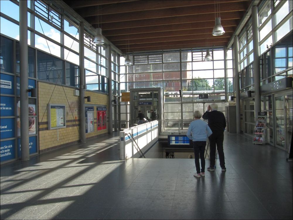 Blick in die Halle eines Bahnhofs. Sie ist bis auf ein älteres (Ehe-?)Paar, das wir von hinten sehen, auf den ersten Blick menschenleer. Die beiden verdecken teilweise die Treppe, an der auf Fußhöhe des Hallenfußbodens eine Abfahrtstafel hängt. Rechts und links je ein Laden, links ferner Fahrplan und Automaten. Zwischen denen und der Treppe der Weg zum zu sehenden Aufzug. Wir sehen auf drei Seiten die Fenster der Halle, hinter Aufzug und Treppe ist ein hinter den nicht sichtbaren Gleisen stehender alter Backsteinspeicher zu sehen.