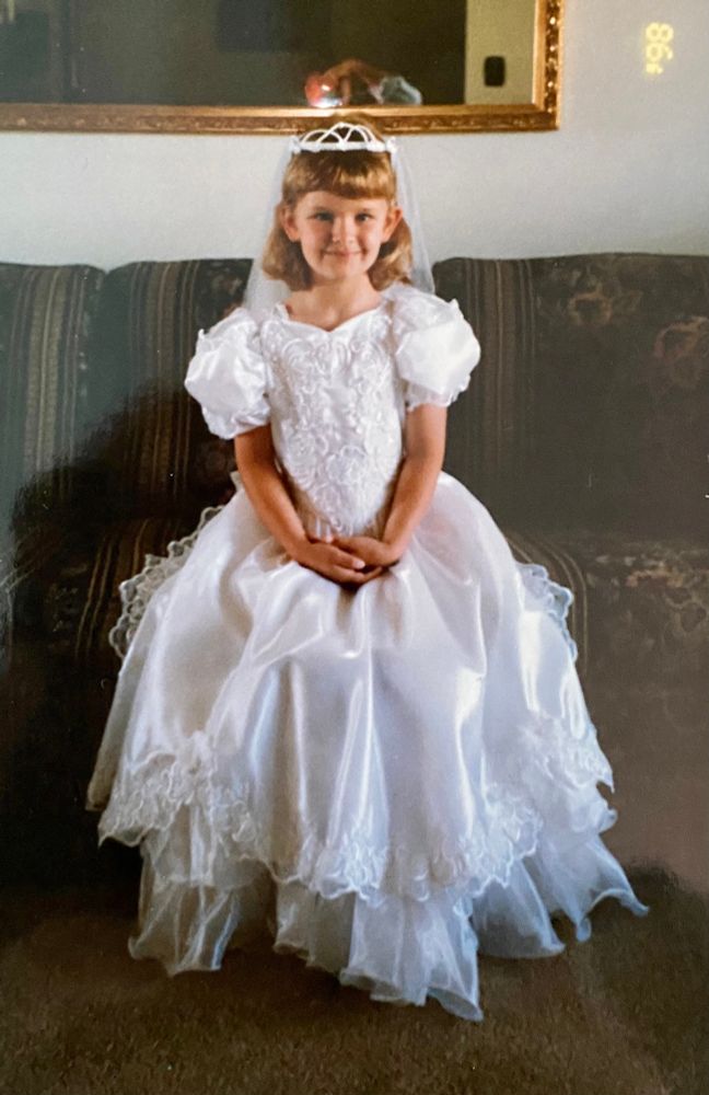 A joyless blonde child in a first communion dress attempts a smile for the camera