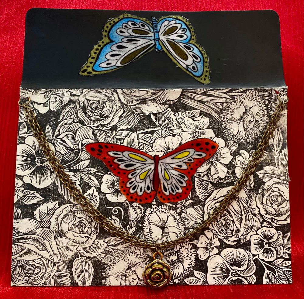 A floral print envelope with a red butterfly in the center, a gold chain with a rose shaped charm, and a blue butterfly on the envelope flap 