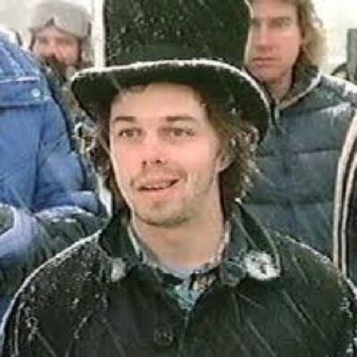 Curtis Armstrong, in character as Charles Demar, wearing a top hat, wool coat, smiling while getting snowed upon.