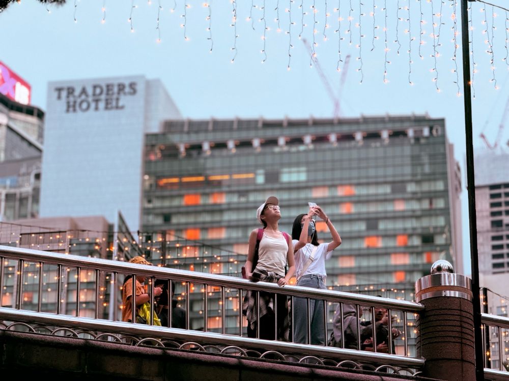 Two women on a Christmas light lit bridge taking a photo of a building in the distance 