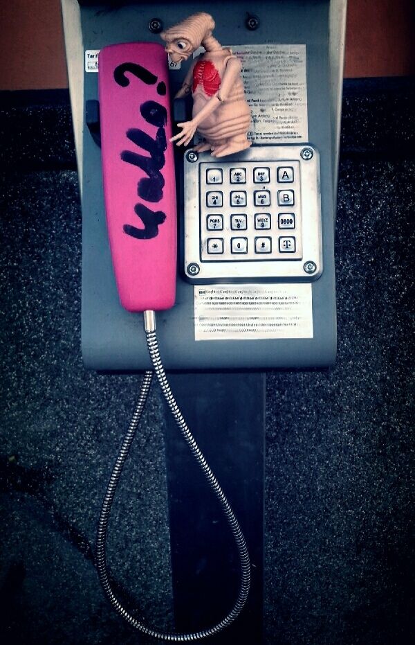 E.T. an einem Münzfernsprecher, auf dem Hörer steht mit Edding "Hallo?" geschrieben 