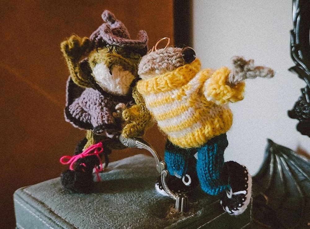 Two handmade crochet toys are lit by the sunlight, one is a frog in a grey witch's dress with glasses and peircings, the other is a toad with golden spectacles, a yellow striped sweater and blue jeans. They are holding hands and posed as if to say "we're going on an adventure!"
