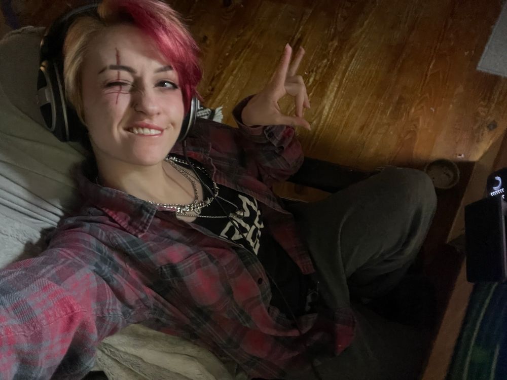 Nox sat in a desk chair with sweatpants and a flannel on over a tank top. They have an upside down cross over their winking eye and the metal horns symbol 