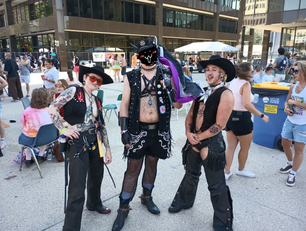In the fourth photo, the same central figure appears again, now posing with two other individuals in front of a large public building during the same Pride event. The person on the left wears a black cowboy hat, red-tinted sunglasses, and an open vest showing off a tattoo-covered chest and arms. Their outfit features lots of accessories, including keys, tools, and fringe details, with long black pants. The person on the right is shirtless, sporting only an open leather vest and a pair of dramatic black chaps over a thong, showing off leg tattoos and body hair. All three appear joyful, expressive, and self-assured, surrounded by fellow festival attendees in casual summer clothing. The photo captures a spirited celebration of identity, creativity, and community at a queer gathering in Grand Rapids, Michigan.