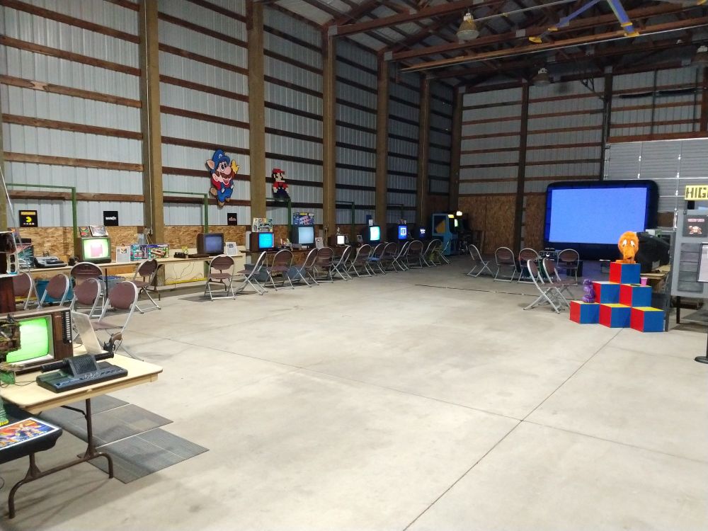 A row of game consoles set up on CRT TVs, a big screen projector, and life-size plus Q*Bert on a red, blue, and yellow pyramid of cubes.