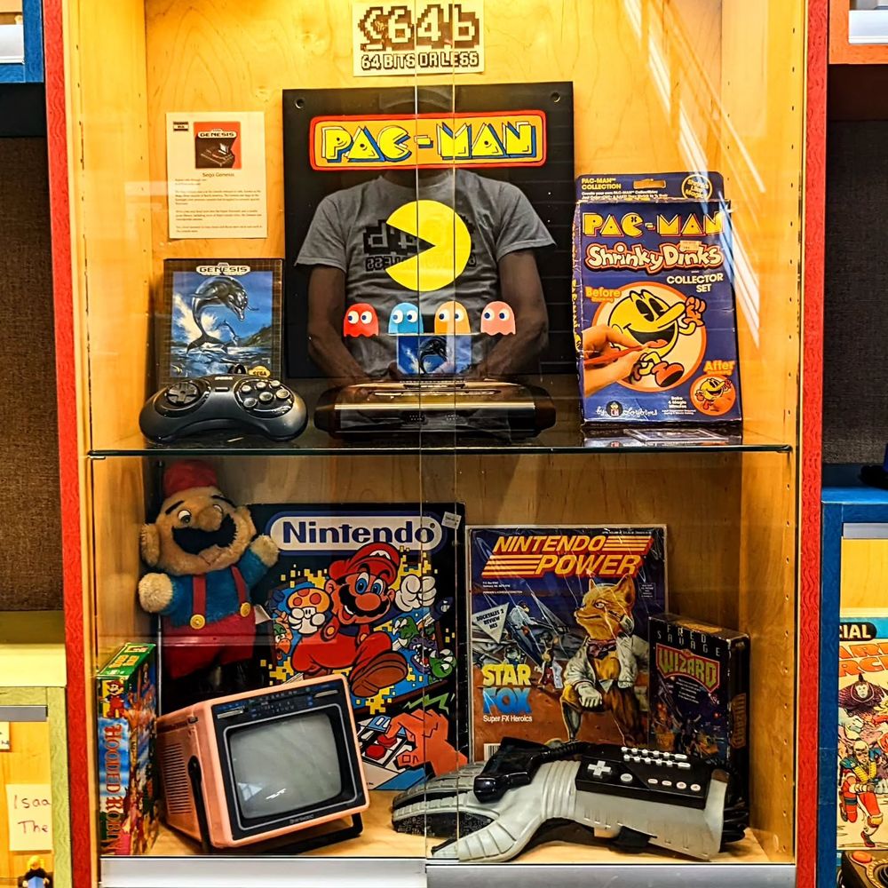 The first case has a Sega Genesis and controller on the top shelf, along with some Pac-Man artwork. On the bottom shelf is a Nintendo power glove, a VHS copy of The Wizard, a Nintendo Power magazine featuring Star Fox, some Mario artwork, and a portable CRT TV set.
