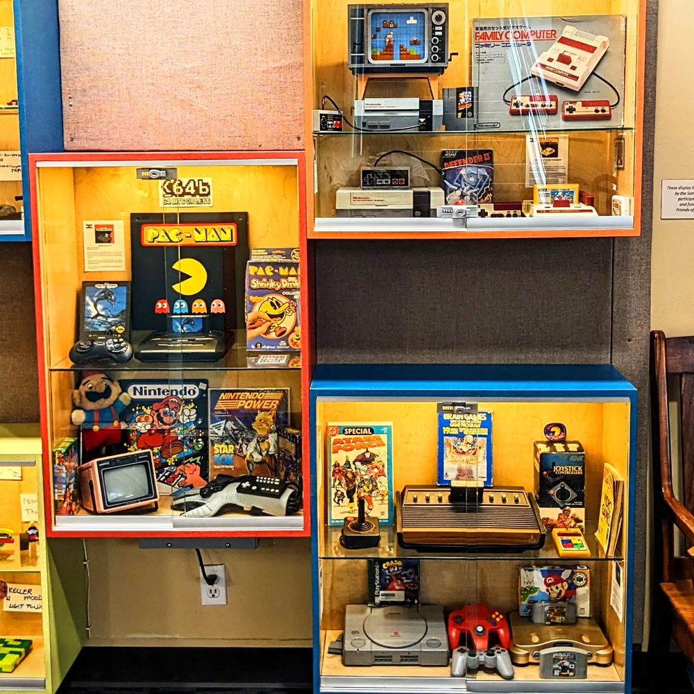 Three glass-front display cases filled with retro gaming consoles, controllers, and memorabilia.