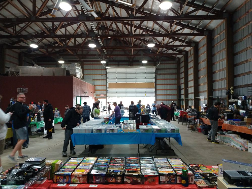 A view of the 64 Bits or Less vendor hall. In the foreground is a table filled with VHS tapes, in the background are several other tables with retro games.