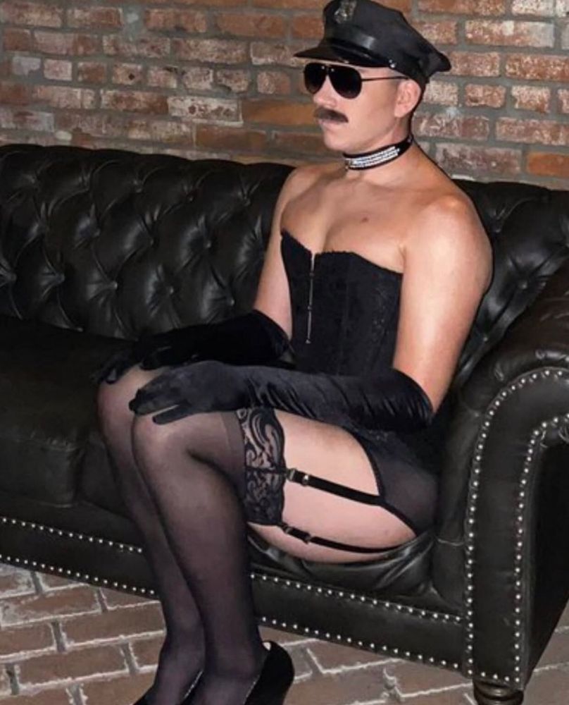 Trixie mattel com chapéu de policial, óculos de sol aviador, bigode grosso, choker de strass, corset preto, luvas de veludo até o cotovelo, cinta liga, meias 7/8 e salto alto. Tudo na cor preta