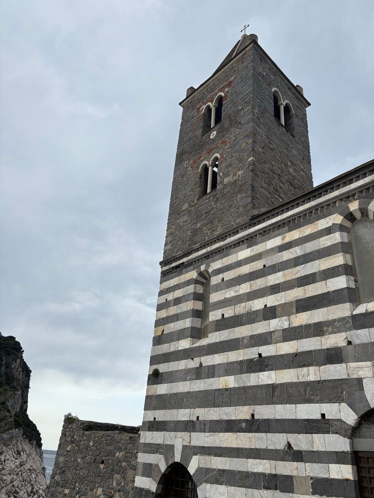 Chiesa di San Pietro, a church at the top of the hill. The main walls are made of alternating dark gray rock and white marble making horizontal stripes from the ground to the roof. The small bell tower above is constructed with smaller dark gray rocks. There is a thin metal cross atop that, dark against the gray cloudy sky. 