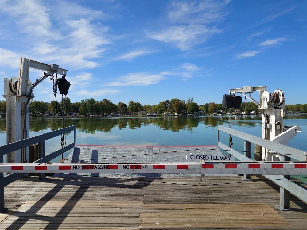 The ramp for the Bemus Point to Stow NY ferry in Bemus Point. A short wooden dock leads to raised metal ramp behind a white and red gate. A sign on the gate reads: CLOSED TILL MAY. Across the narrowest section of Chautauqua Lake, the matching raised ramp and dock on the Stow, NY side.