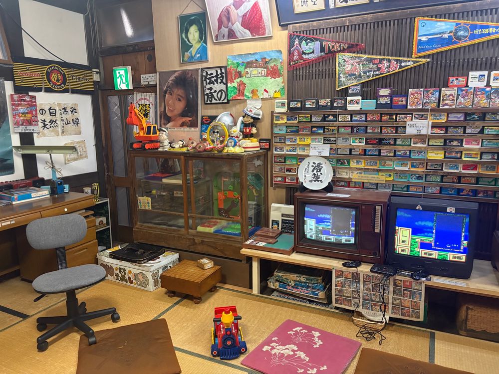 Ein Foto von einer Nachstellung eines japanischen Wohnzimmers im Showa Museum von Takayama. Man sieht mehrere flache Kissen auf Tatamimatten vor zwei kleinen kastigen Fernsehern. Auf der Wand dahinter sind Dutzende Retrospiele angereiht. 