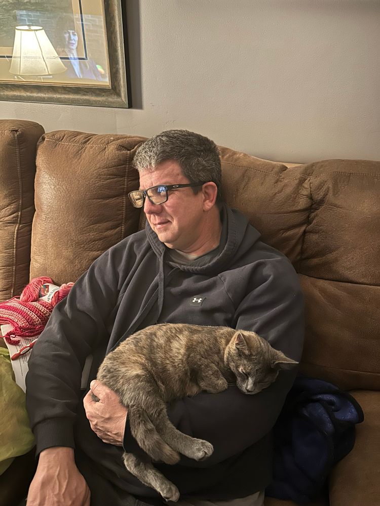 A man with dark hair sits on a brown sofa cradling a sleeping dilute tortie kitten