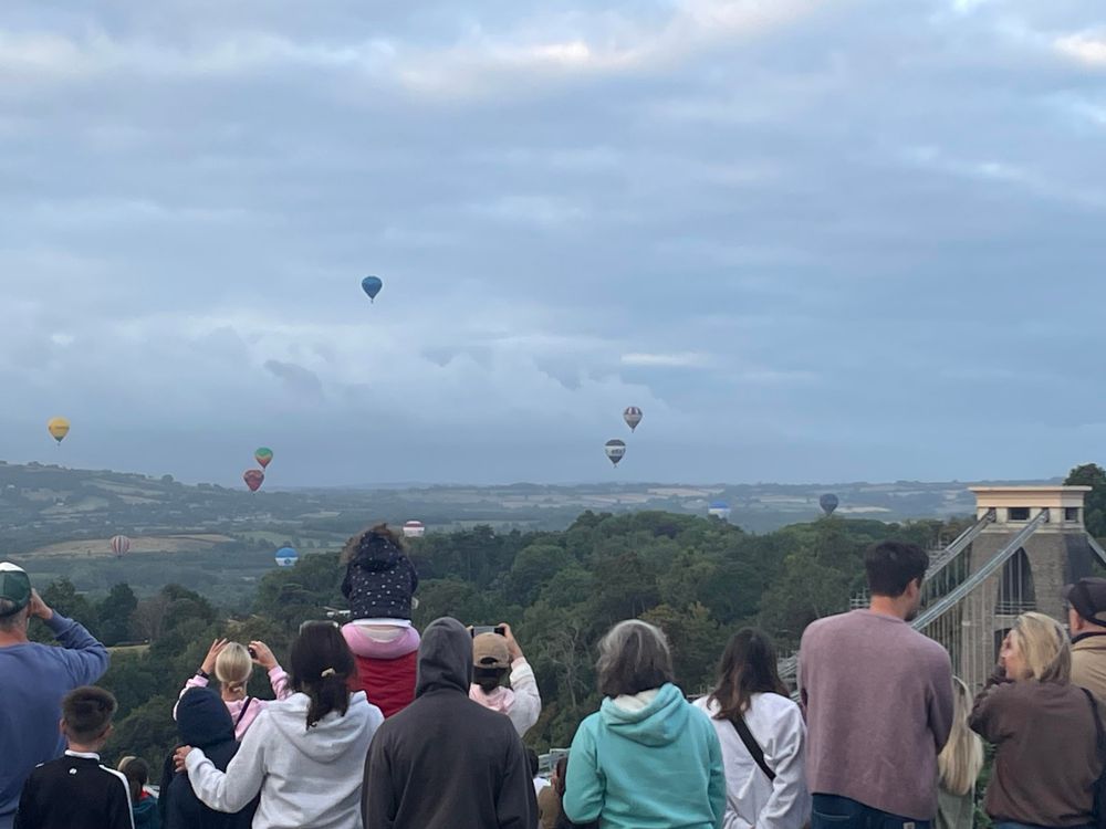 A crowd of people watching multiple hot air balloons floating over the Clifton suspension bridge.